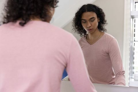 Non-binary person standing at vanity mirror in pink v-neck reflecting morning routine