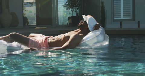 Muscular man lounging on clear pool float wearing pink trunks and visor, summer lifestyle