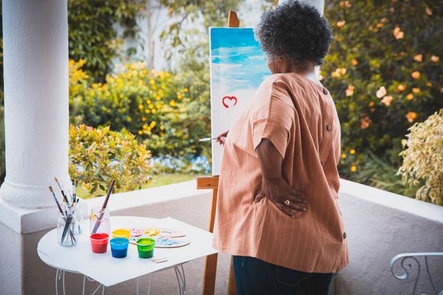 African american senior woman painting on canvas on terrace in sunny garden. staying at home in isolation during quarantine lockdown.
