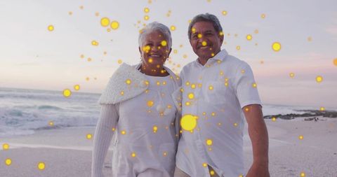 Senior couple walking on beach at sunset, smiling and embracing amid glowing golden bokeh