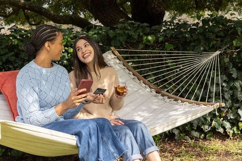 Diverse women relaxing on backyard hammock, sharing smartphone and credit card, smiling