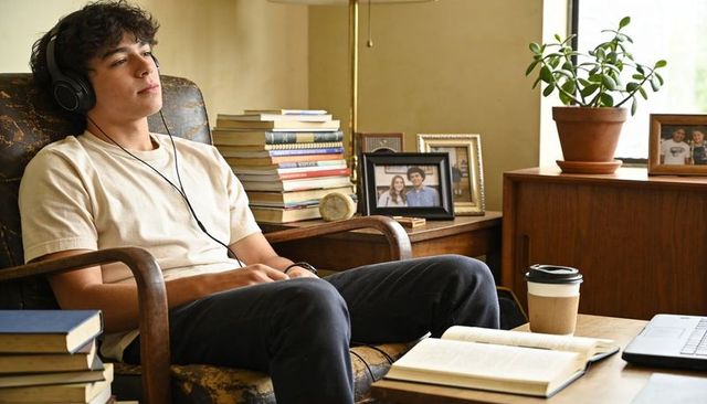 Teen listening on over-ear headphones relaxing with books and laptop in cozy armchair