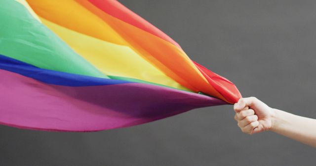 Hand holding rainbow Pride flag against gray background. Represents LGBTQ pride, equality, and support. Perfect for using in campaigns, social media, LGBTQ events, awareness, promotional materials, and articles about diversity and inclusion.
