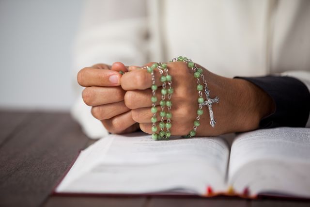 Woman's hands holding a green rosary while praying over an open Bible on a wooden desk. Ideal for use in religious publications, faith-based websites, spiritual blogs, and educational materials on prayer and devotion.