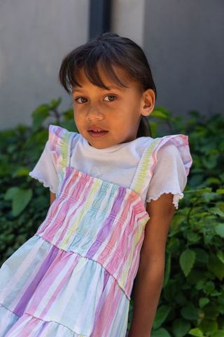 Young girl wearing pastel striped sundress and white ribbed top standing in sunlit garden