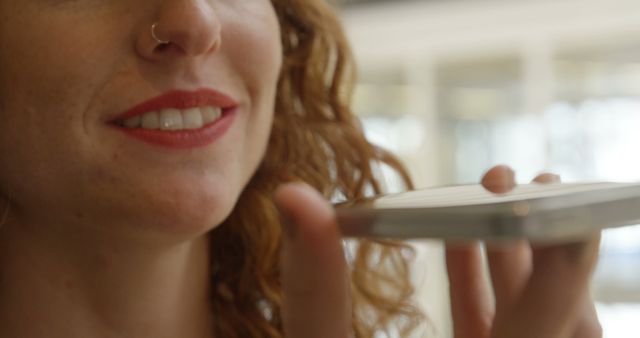 Woman with red lips and curly hair using a voice command feature on her smartphone. Close-up view focuses on the smile and phone interaction. Great for depicting modern technology usage, communication, and voice assistant features in everyday life.