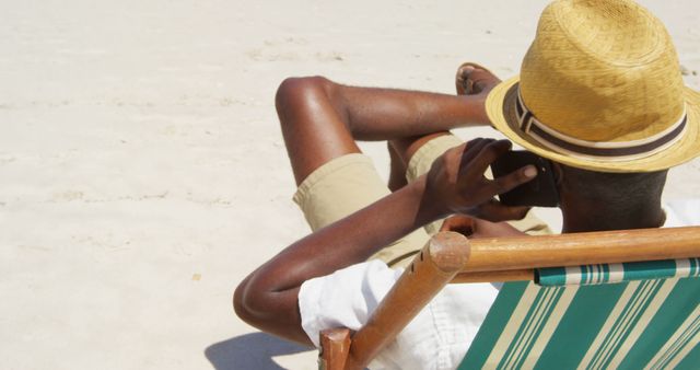 A middle-aged African American man relaxes on a beach chair, wearing a straw hat and using a smartphone, with copy space. His leisurely pose suggests a moment of tranquility during a sunny beach vacation.