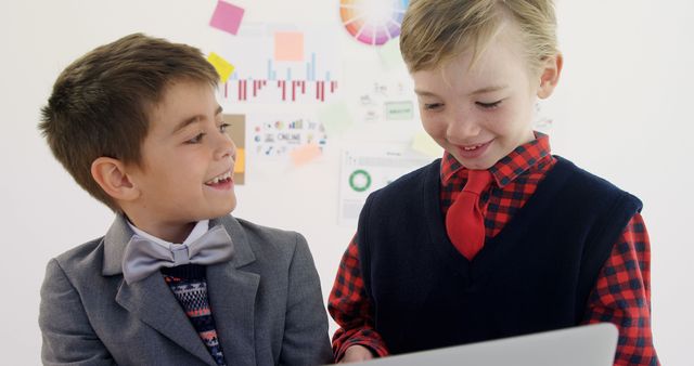 Two young boys in formal attire collaborating on a laptop in a brightly lit classroom. They are both engaged and happy while working together. This could be used for educational materials, promoting teamwork and learning in technology environment, and highlighting children in modern educational settings.