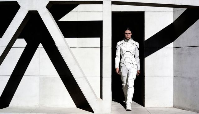 Woman in futuristic white exosuit stepping from dark doorway amid geometric concrete