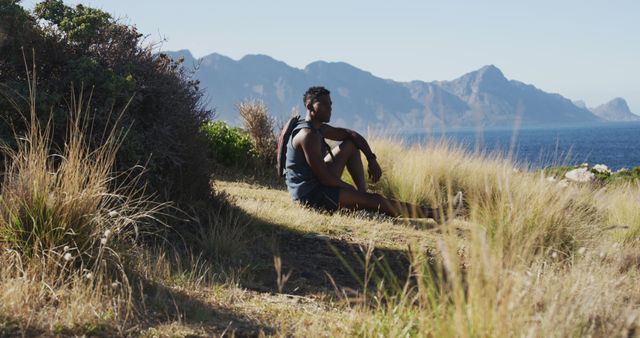 Young man resting on a grassy hillside with a picturesque view of mountains and sea in the background, suitable for themes of travel, adventure, outdoor activities, nature exploration, and relaxation. Perfect for promoting tourist destinations, travel blogs, adventure lifestyle content, and outdoor gear.