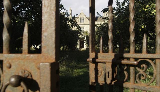 Framing Rusted Iron Gate Leading Through Overgrown Orchard Toward Stone Manor