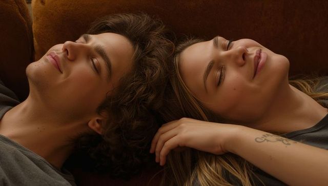 Young couple resting on brown couch in warm cozy moment, forearm tattoo, intimate calm