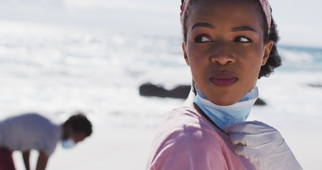 This image shows a young woman wearing gloves and adjusting her face mask while standing on a beach during the day. She is looking away pensively as another person can be seen in the background wearing a face mask. This photo can be used for topics related to environmental conservation, beach cleanups, personal safety during the pandemic, outdoor activities, or volunteer work.