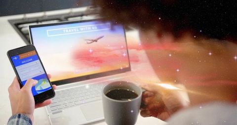Checking travel app on smartphone holding coffee by laptop, home desk nomad lifestyle