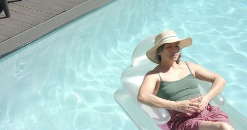 Senior asian woman relaxing on inflatable pool float, wearing straw sun hat and sipping drink