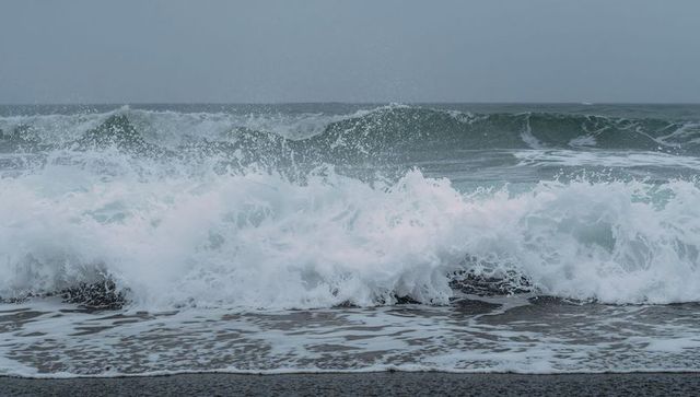 Crashing ocean waves creating foamy surf on dark pebble beach under overcast sky