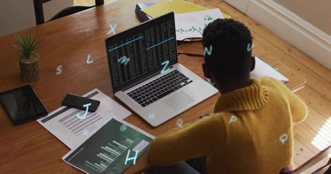 Typing woman in mustard sweater analyzing bar charts while coding on laptop at home