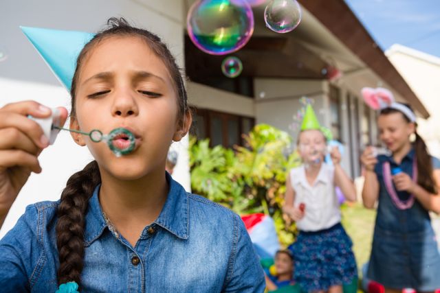 Girl blowing bubbles while standing in yard during an outdoor party. Other children in the background wearing party hats and enjoying the celebration. Ideal for use in advertisements, blogs, or articles about children's parties, outdoor activities, and summer fun.