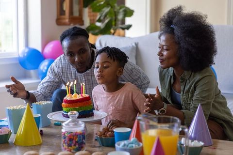 African american family celebrating child birthday blowing candles on rainbow cake at home
