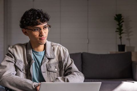 Young asian man working from home on laptop wearing glasses and denim jacket on couch