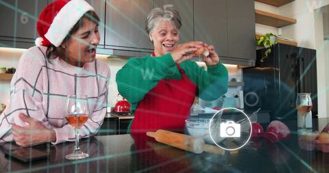 Senior woman cracking egg while friend in santa hat laughing during holiday baking in cozy kitchen