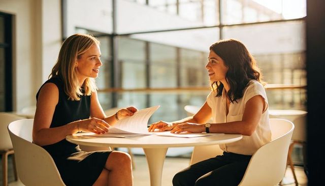 Businesswomen reviewing documents in sunlit office atrium during collaborative meeting