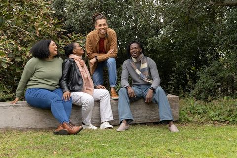 Multiracial friends laughing on wooden log bench in autumn park, casual outdoor gathering