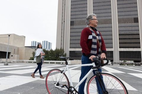 Senior man with bicycle pausing on urban plaza while young woman walking behind him