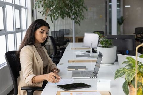 Indian businesswoman working at laptop in modern open-plan office with plants and notepad