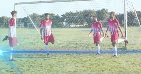 Four soccer players stretching quadriceps in red jerseys by goal on sunlit grass field