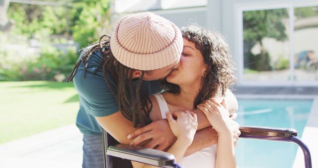Happy biracial woman in wheelchair embracing and kissing with male partner with dreadlocks in garden. wellbeing and domestic lifestyle with physical disability.