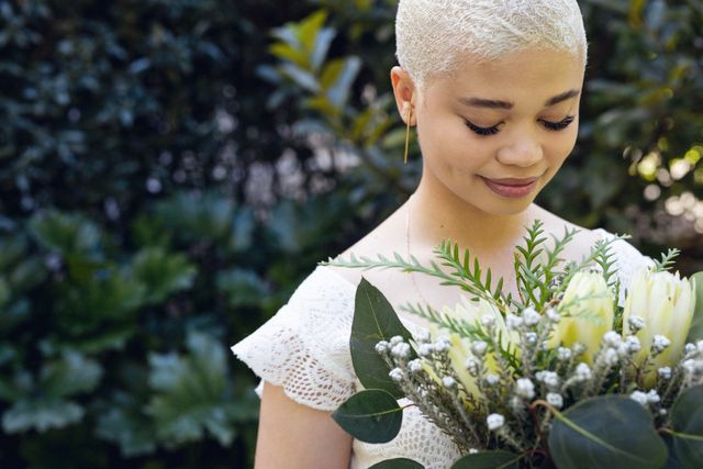 Close-up of smiling biracial young woman with short hair looking at flowers against plants in yard. Unaltered, happy, lifestyle and nature concept.