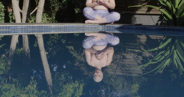 The image shows a woman meditating by a swimming pool with her reflection visible in the water. She is wearing yoga pants and is in a serene outdoor environment surrounded by greenery and trees. This image can be used to illustrate themes related to mindfulness, peace, tranquility, yoga, and wellness.