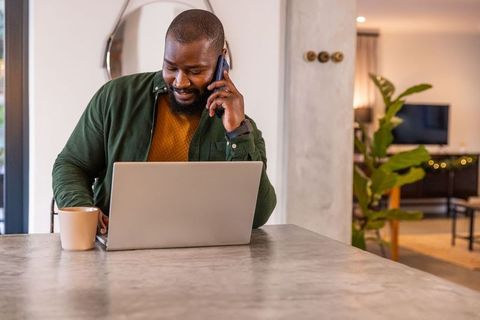 African american man working remotely on laptop while talking on phone at home