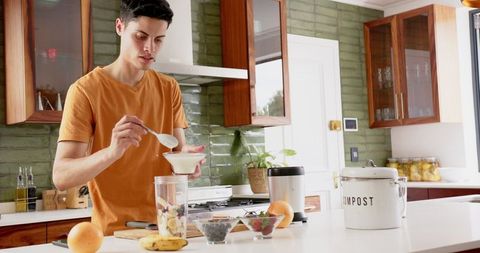 man adding yogurt to a fruit smoothie with fresh ingredients in a stylish kitchen. useful for promoting healthy lifestyles, nutrition blog content, or advertising kitchen appliances. highlights positive home cooking and wellness choices.