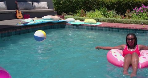 Sunlit backyard pool relaxation: african american woman floating on pink donut float, smiling