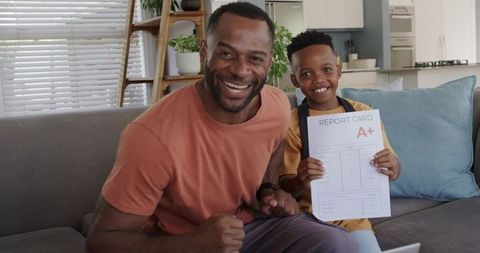 African american father celebrating son a+ report card on couch in modern home