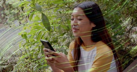 Asian woman texting on smartphone sitting in leafy garden with light streak overlays