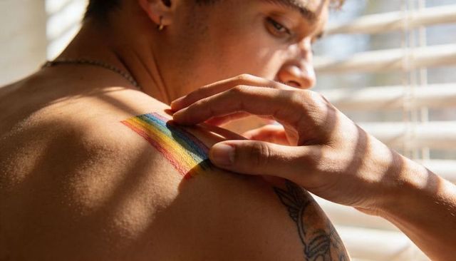 Shirtless person touching rainbow shoulder paint basking in warm window light