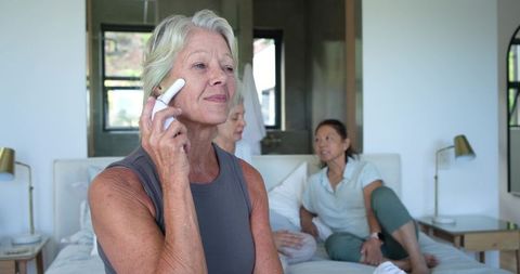 Using handheld facial device by mature woman while diverse friends relaxing on cozy bed