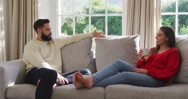Couple sitting on sofa, engaging in relaxed conversation. Man gestures while talking; woman listens, holding coffee cup. Ideal for use in relationship articles, home decor advertisements, and lifestyle blogs showcasing comfortable family settings or effective communication.
