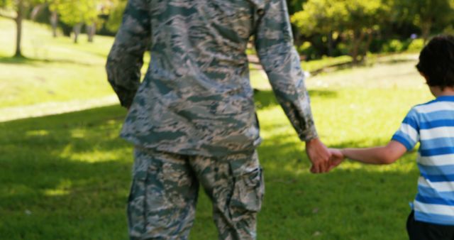 Depicts military parent and child holding hands and walking in lush, green park. Great for family and bonding concepts, military family campaigns, parenting blogs, and promotional material about military support or family time.