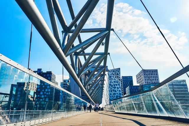 This image displays a modern pedestrian bridge with striking architectural elements in an urban setting. The bright sunny day casts a clear view on the cityscape with glass buildings and steel structures reflecting modernity. This photo is ideal for illustrating concepts of modern architecture, urban development, contemporary city life, or environmental design in presentations, advertisements, and editorial content.