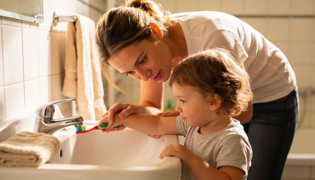 Mother guiding toddler brushing teeth at sink, teaching dental care and morning routine