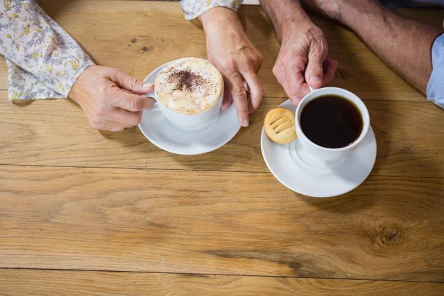 Senior couple enjoying coffee together at a wooden table. One person has a cappuccino, and the other has a black coffee with a biscuit. Ideal for use in articles or advertisements about senior lifestyle, coffee culture, or social activities for the elderly.