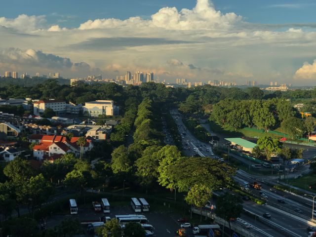 Aerial view capturing an urban landscape featuring a busy highway surrounded by lush trees and green spaces. The image shows the contrast between nature and urban development, with the city's skyline visible in the distance beneath a sky filled with clouds. Ideal for use in real estate promotions, urban planning presentations, or travel and tourism marketing materials highlighting cityscapes and urban design.