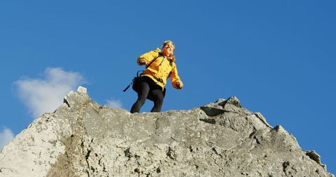 Captured is a woman standing triumphantly on a rocky mountain summit under a clear blue sky. Her yellow jacket contrasts starkly against the rock face and makes her the central figure of this image, epitomizing success and exploration spirit. Perfect for use in articles or advertisements focused on adventure tourism, motivational themes, and outdoor sports.