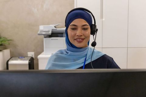 African-american woman in blue hijab smiling as call center agent with headset at desk