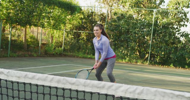 A young woman is playing tennis on an outdoor court surrounded by trees and green foliage. She is dressed in sports attire and holding a tennis racquet, preparing to hit the ball. This image conveys a sense of fitness, healthy lifestyle, and sporting activity. It is ideal for use in advertisements and promotional material for sports equipment, fitness programs, activewear brands, and recreational facilities.