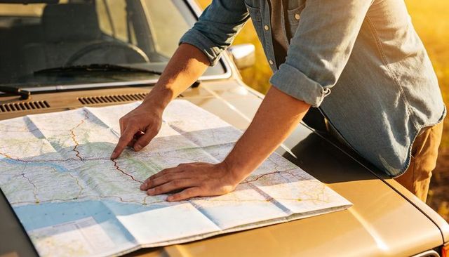 Leaning traveler studying paper map on car hood at sunset planning road trip route